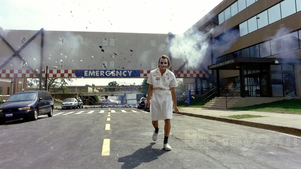 A man in a hospital gown wearing smudged clown make up walks away from a hospital explostion