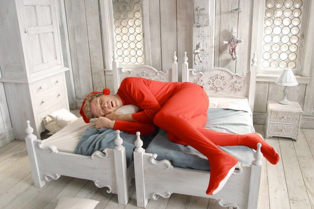 A middle aged man, Will Ferrell, lies across two tiny beds dressed as an elf in a red jumpsuit.
