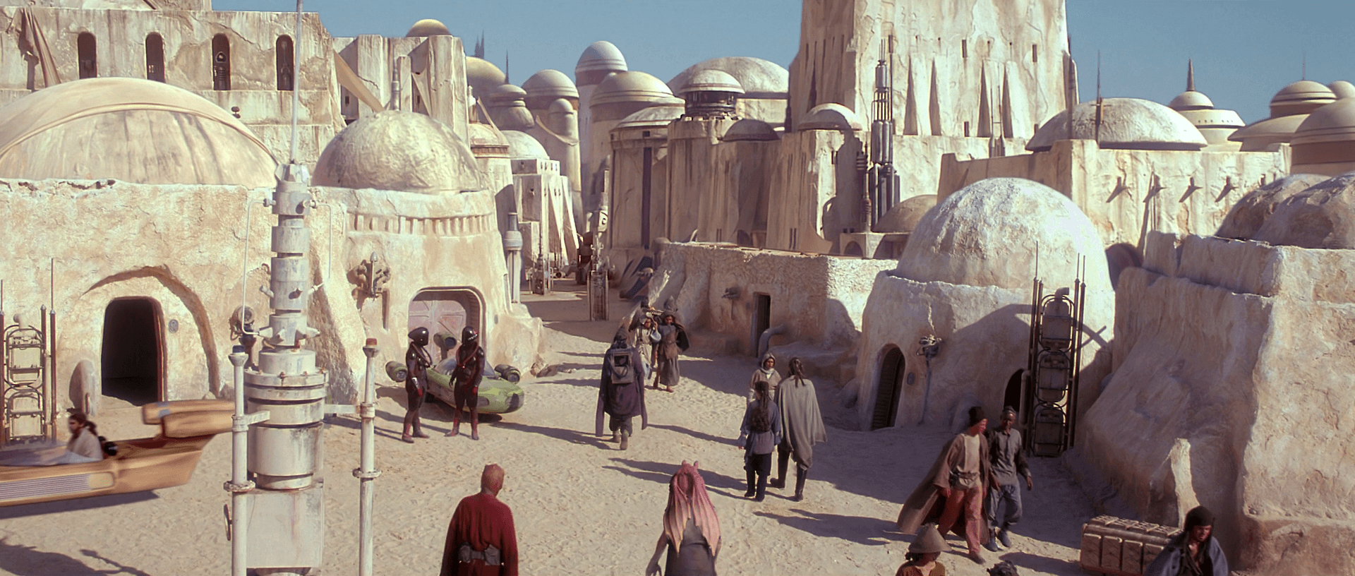 The fictional city of Mos Espa is located within the sand dunes of Tunisia. The characters from Star Wars are walking down the marketplace with many domed buildings and sci fi structures around them. 