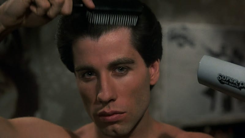 Image featured is a still from the movie Saturday Night Fever. Pictured is John Trovolta combing and blow drying his hair in the mirror.